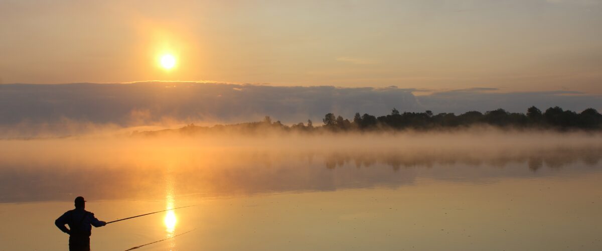 Ein Angler steht im Wasser im Morgengrauen  Im Hintergrund die aufgehende Sonne 