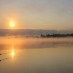 Ein Angler steht im Wasser im Morgengrauen  Im Hintergrund die aufgehende Sonne 