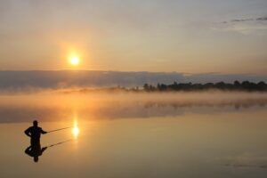 Ein Angler steht im Wasser im Morgengrauen  Im Hintergrund die aufgehende Sonne 
