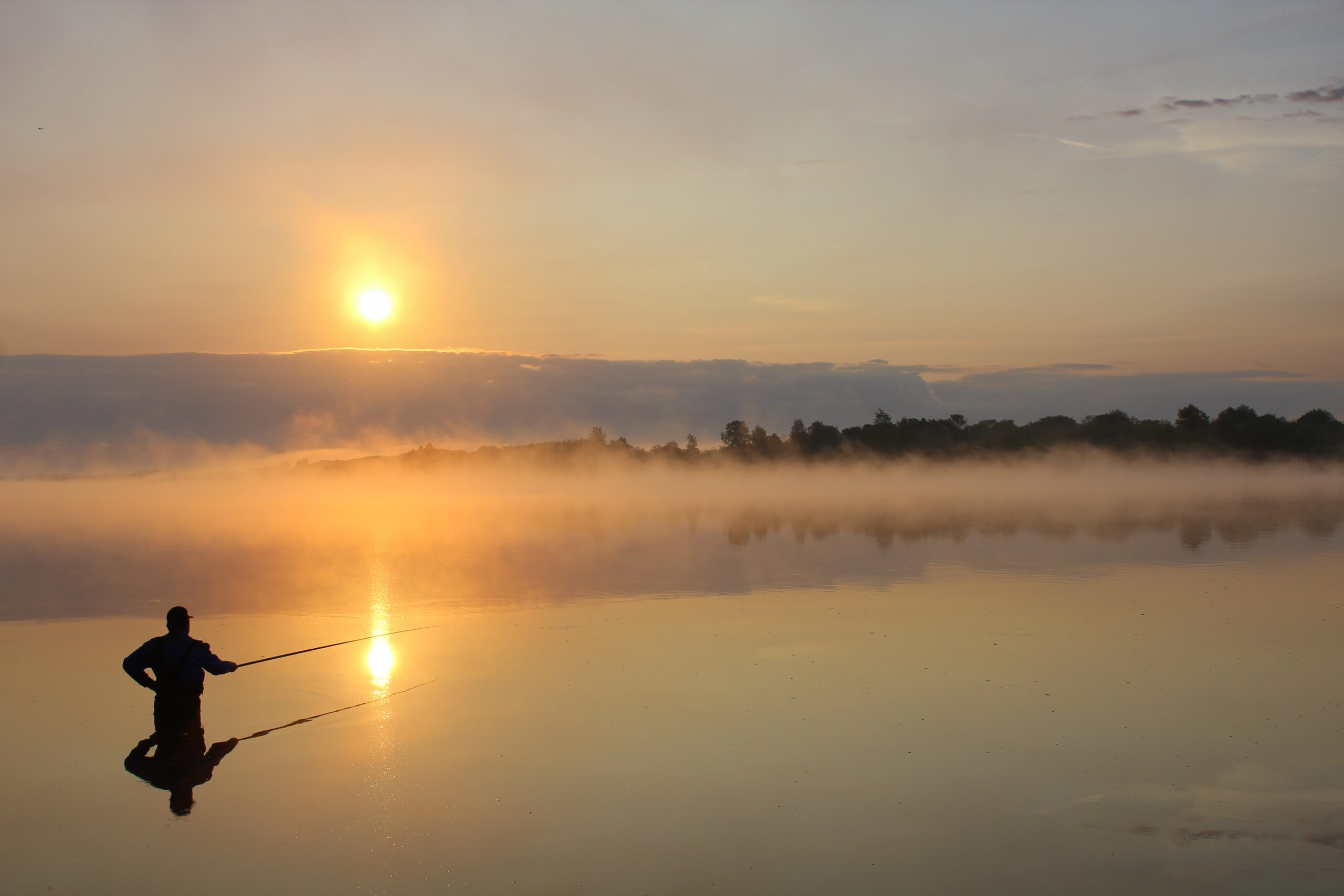 Ein Angler steht im Wasser im Morgengrauen  Im Hintergrund die aufgehende Sonne 